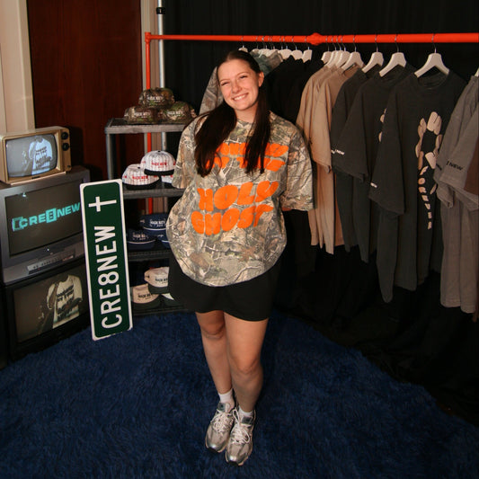 model wearing a sponsored by the Holy Ghost christian faith-based shirt camouflage outfit with orange accents in a room with multiple television screens displaying 'CRE8NEW'.